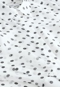 Chemise blanche boutonnée avec des pois noirs et gris, dotée d’un col classique et de boutons sur le devant.
