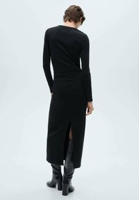 Robe noire à manches longues avec une silhouette ajustée, fente latérale et texture douce, associée à des bottes noires montantes.