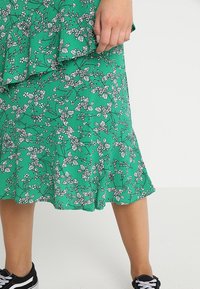 Green floral midi skirt with ruffle detail worn with black sneakers, showing lower legs and one hand resting on the skirt.
