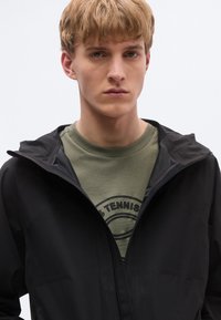 Fekete cipzáras kapucnis dzseki matt felülettel. Alatta olívazöld pulóver, kör alakú tenisz grafikával.