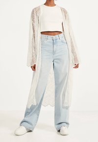 Person som bär högmidjade ljusblå vida jeans, vit crop top, genomskinlig vit lång cardigan med blommönster och vita sneakers.