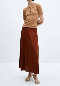 Persona vistiendo una camiseta de polo beige de mangas cortas con relieve, una falda maxi plisada color óxido y sandalias de tacón blancas, de pie contra un fondo blanco.
