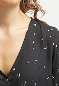 Blouse noire en tissu léger avec un col en V, ornée de petits motifs d'oiseaux blancs sur toute la surface.