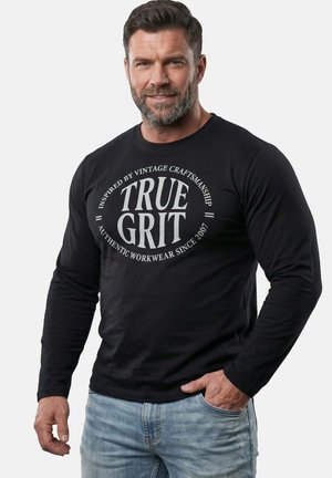 Mężczyzna z brodą w czarnej koszuli z długim rękawem z napisem "TRUE GRIT" i jasnoniebieskich dżinsach, stojący z jedną ręką w kieszeni.