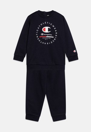 Tuta a due pezzi blu navy realizzata in tessuto morbido. Presenta maniche lunghe, polsini elasticizzati e un grande logo sulla parte anteriore.