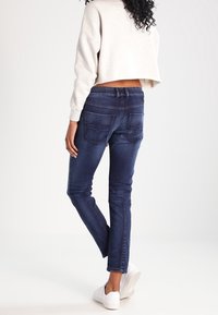 Jeans azul oscuro de corte ajustado con bolsillos traseros, combinados con un sudadera cortada de color gris claro. El modelo lleva zapatillas blancas.