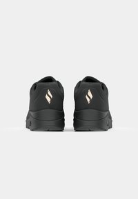 Paire de baskets noires vues de dos avec un logo clair en forme de losange sur chaque talon.