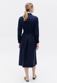 Abito lungo blu navy con maniche lunghe, colletto, vita aderente e spacchi laterali. Realizzato in un tessuto testurizzato, con dettagli a bottoni sulle maniche.