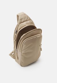 Beige ryggsäck med vadderat tyg, flera dragkedjefack och en unik triangulär form. Har en mjuk textur och robusta detaljer.