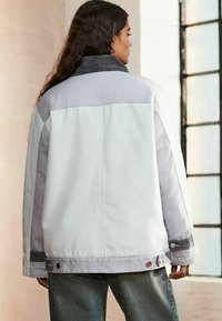 Witte en grijze spijkerjack met een hoge kraag, knoopdetails en contrasterende donkergrijze accenten op de schouders en manchetten.