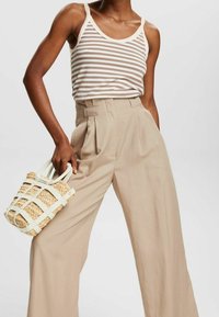 Mujer con pantalones de tiro alto y pierna ancha en color beige, una camiseta sin mangas a rayas blancas y beige, sosteniendo un bolso de mano de mimbre con asas blancas.