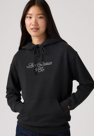 Sudadera negra hecha de una tela suave, con un bolsillo frontal, capucha con cordones y un logo bordado en blanco. Textura de punto de jersey.