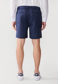 Shorts bleu marine en tissu léger, avec une taille décontractée, des revers roulés et deux poches arrière. Portés avec des baskets blanches.