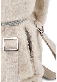 Acercamiento a una mochila de piel sintética beige con un bolsillo lateral sin cerrar y correas de tela beige ajustables con hebillas plateadas.