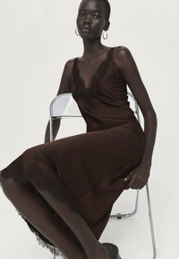 Bruine satijnen slipjurk met kanten bies langs de halslijn en zoom, met een aansluitend silhouet en verstelbare bandjes, gedragen met een stoel als achtergrond.