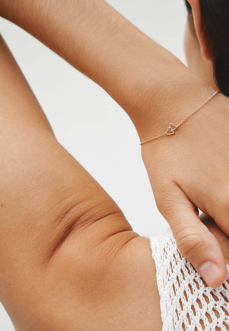 Pulsera de oro con un pequeño encanto en forma de corazón, exhibida en una muñeca de piel marrón clara. La cadena de la pulsera es delicada y fina.