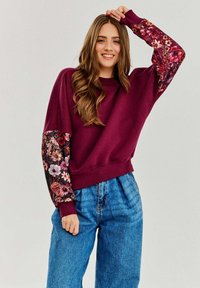 Bordowy sweter z rękawami w kwiatowy wzór, okrągłym dekoltem i luźnym krojem, zestawiony z niebieskimi spodniami dżinsowymi o wysokim stanie.