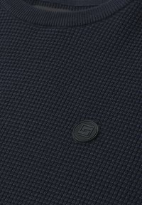 Pull tricot gaufré bleu foncé avec un col rond et un petit patch rond noir avec logo sur la poitrine.