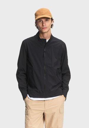 Jeune homme portant une casquette beige, une veste noire zippée, un t-shirt blanc en dessous et un pantalon cargo beige, debout les mains dans les poches.