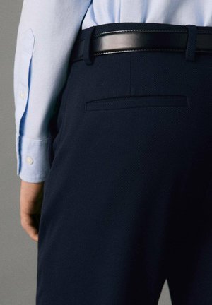 Pantalones navy con una tela texturizada, que presentan un bolsillo ribeteado y un cinturón de cuero negro. Una camisa azul clara es parcialmente visible en la parte superior.