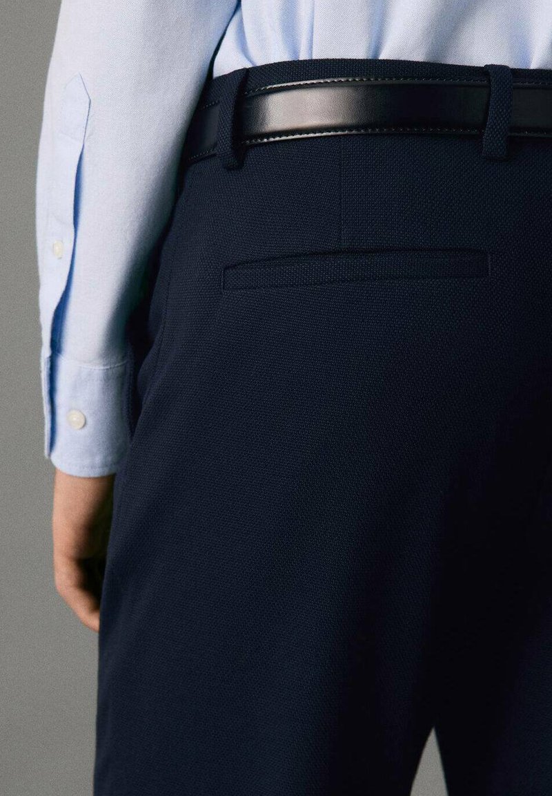 Námornícke nohavice s textúrovanou látkou, s priečnou kapsou a čiernym koženým opaskom. Svetlomodrá košeľa je čiastočne viditeľná nad nimi.