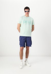 Light green cotton t-shirt with "GUESS" graphic, paired with navy blue cargo shorts and white sneakers for a casual look.