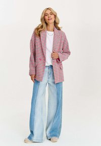 Blonďatá žena v růžovém kostkovaném saku, bílém triku, širokých dvoubarevných modrých džínách a béžových botách, stojící proti bílé zdi.