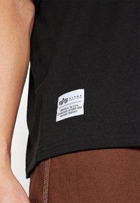 Sort T-shirt lavet af bomuld med et hvidt mærke, der har logo og tekst, med kantede kanter, vist sammen med brune bukser med synlig syning.