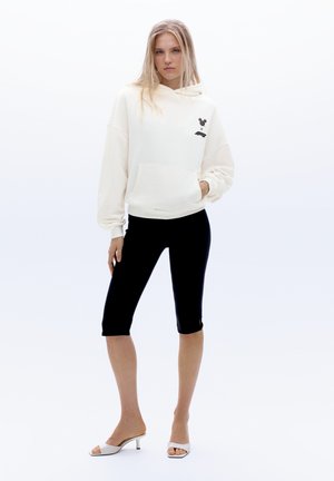 Femme portant un sweat à capuche blanc avec un petit logo noir, des leggings courts noirs et des talons blancs à bout ouvert, debout devant un fond blanc uni.