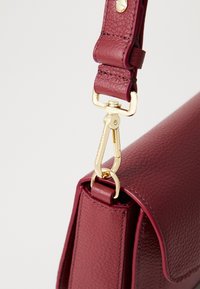 Close-up of maroon leather bag strap attached with gold-toned clasp and hardware against plain light background.