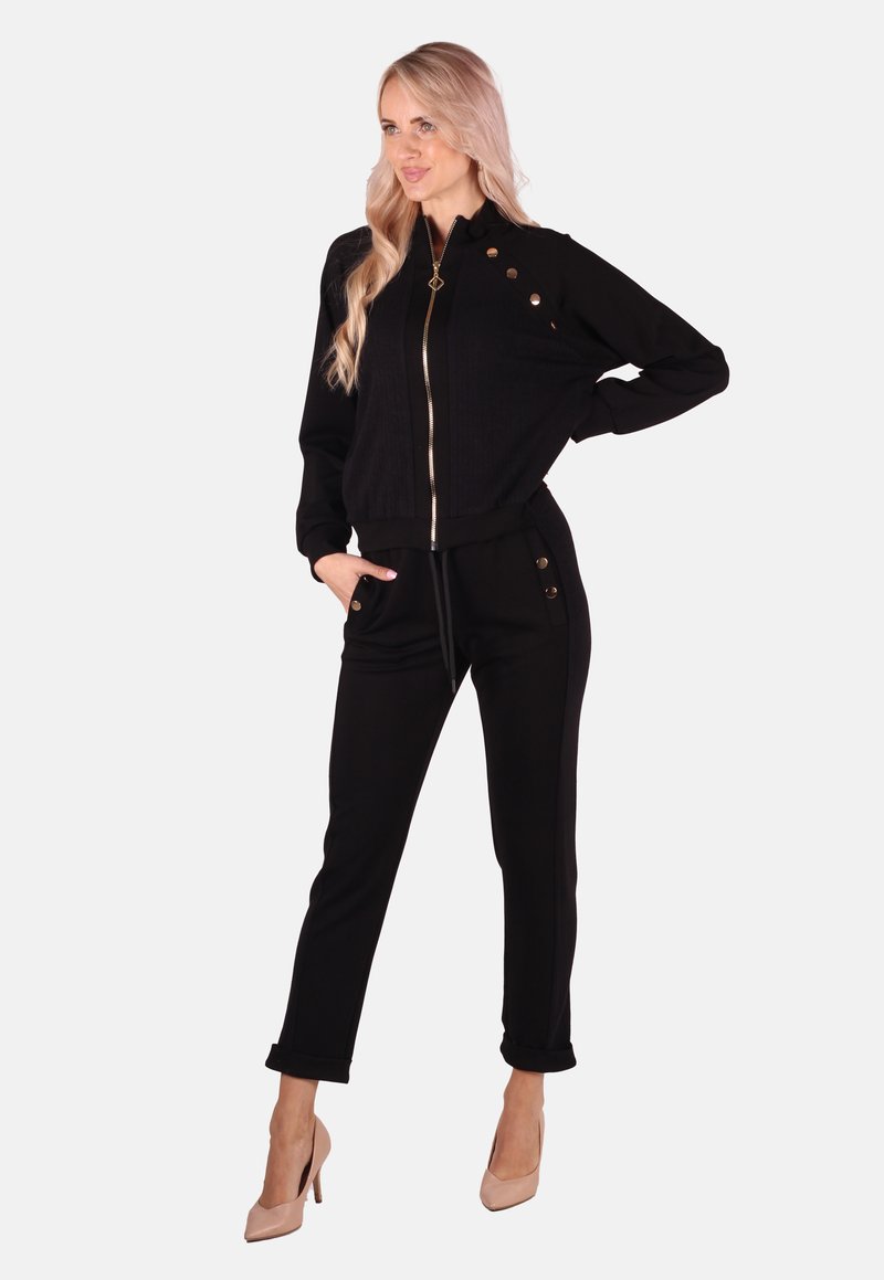 Schwarze Zip-Jacke mit gerippter Textur und metallischen Knopfdetails, kombiniert mit schwarzen Hosen mit seitlichen Knöpfen und schmaler Passform.