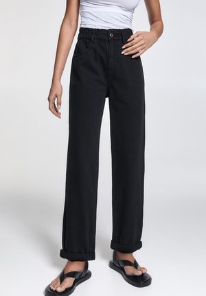 Femme portant un pantalon noir taille haute à jambes larges avec revers retroussés, un top blanc sans manches et des sandales plateformes noires sur un fond uni.