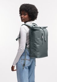 Grå rull-top ryggsäck med justerbara remmar, texturerat material, framficköppning med dragkedja och minimalistisk design, buren av en person mot en neutral bakgrund.