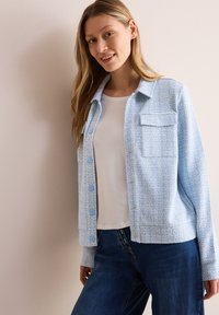 Lichtblauwe, textuurjasje met een knopencommissie aan de voorkant, één borstzak en afgeronde zoom, gedragen over een wit shirt en donkere spijkerbroek.