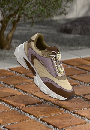Beiger und brauner Sneaker mit lila Akzenten, strukturiertem Mesh und glatten Materialien, ausgestattet mit weißer Sohle und markiertem Schnürsenkelhalter.