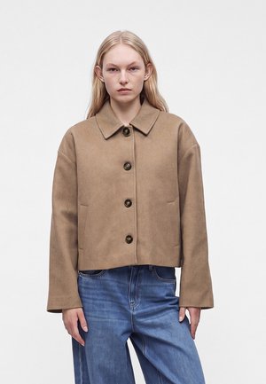 Ung kvinde iført en beige uldjakke med knapper og lange ærmer samt blå jeans, stående foran en ensfarvet hvid baggrund.