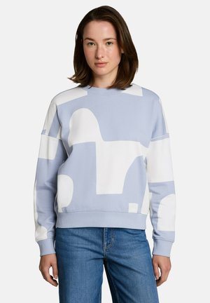 Jonge vrouw met bruin haar tot op de schouders, die een lichtblauw-wit sweatshirt met abstract patroon en blauwe spijkerbroek draagt, staand tegen een witte achtergrond.