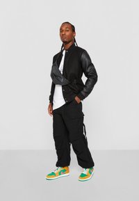 Chaqueta bomber negra con mangas de cuero, camiseta blanca, pantalones cargo negros y zapatillas de colores. Incluye un bolso cruzado negro.