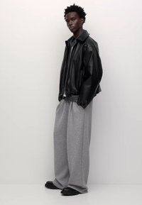 Mladý muž stojící v černé kožené bundě, širokých šedých teplácích a černých botách na pozadí jednoduché bílé stěny.