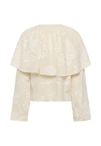 Blouse crème à manches longues avec cape à épaules larges et volants, ornée de broderies florales blanches sur les manches et le dos.