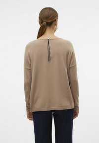 Pull beige à manches longues avec une coupe décontractée, doté d'une fermeture éclair dans le dos et d'une finition texturée. Un jean sombre complète la tenue.