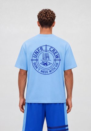 Osoba z krótkimi kręconymi włosami, ubrana w jasnoniebieski t-shirt z ciemnoniebieskim okrągłym logo i tekstem "UNFR CREW DON'T MESS WITH US" na plecach.