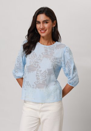 Blusa azzurra chiara in tessuto trasparente, con motivo floreale ricamato, scollatura rotonda e maniche a sbuffo, abbinata a jeans bianchi.