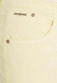 Žluté džíny z denimu mají zadní kapsu s bronzovou štítkem, na kterém je napsáno "cream denim", a tónové švy, které zdůrazňují čistou, hladkou texturu.