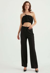 Top corto nero con spalline e design plissettato abbinato a pantaloni palazzo neri con vita alta e plissettati, indossati con décolleté nere a punta.