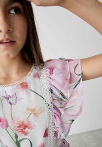 Blumenkleid mit kurzen, pinken, transparenten Ärmeln, weißem Grund, aufwendigem Spitzenbesatz und verschiedenen Blumenmustern in sanften Farben.