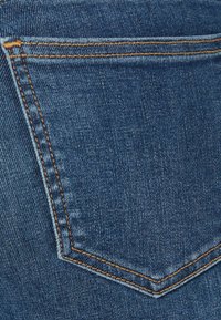 Bolsillo trasero de jeans de mezclilla con tela azul oscuro, costuras visibles en naranja y una textura suave con un ligero brillo.