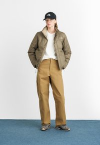 Olivgrüner gepolsterter Mantel, weißer Pullover, braune Hose, schwarze Mütze; verschiedene Texturen mit Reißverschlüssen und Nähdetails. Graue Schuhe runden das Outfit ab.