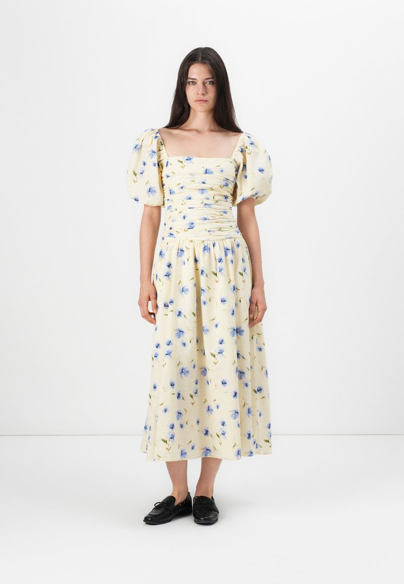 Abito midi floreale con maniche a sbuffo, caratterizzato da una scollatura quadrata, corpino arricciato e un tessuto giallo chiaro con motivi floreali blu. Scarpe nere.