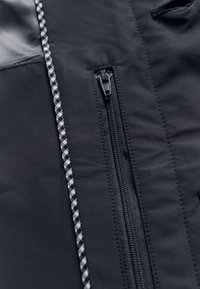 Artimos planas juodos medžiagos striukės su vertikaliu užtrauktuku ir šalia užtrauktuko einančiu juodai balto raštuoto juosteliu.